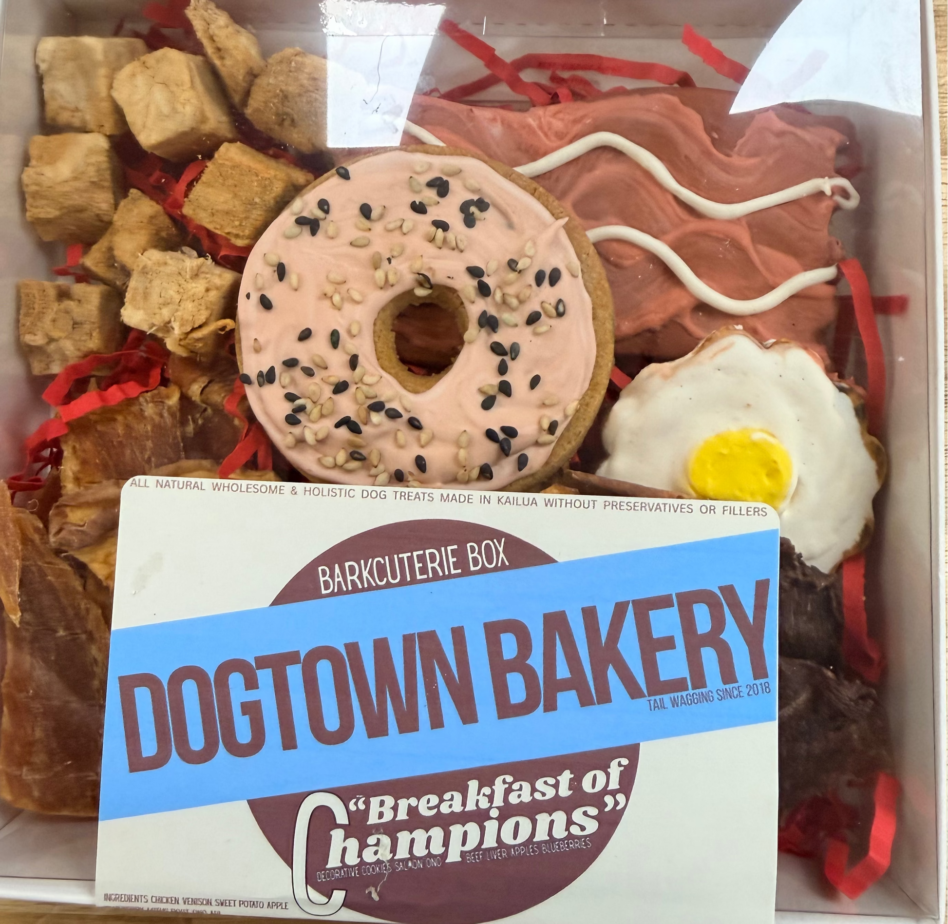 A dog treat box from Dogtown Bakery featuring a pink frosted donut with black sesame seeds, bacon-flavored treats, chicken treats, a fried egg treat, and a white sign in front that reads 'Dogtown Bakery Breakfast of Champions'.