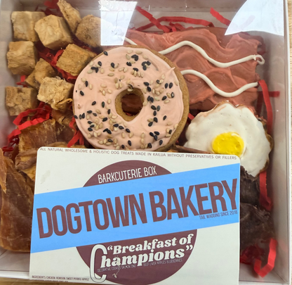 A dog treat box from Dogtown Bakery featuring a pink frosted donut with black sesame seeds, bacon-flavored treats, chicken treats, a fried egg treat, and a white sign in front that reads 'Dogtown Bakery Breakfast of Champions'.